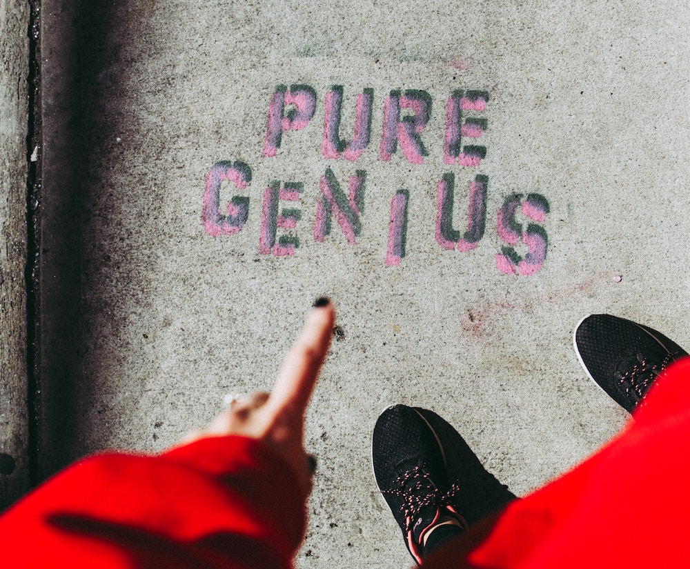 A finger pointing to "Pure Genius" spraypainted on a sidewalk.